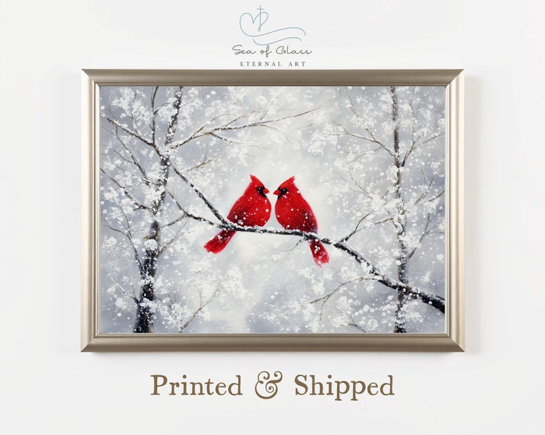 May include: A framed painting depicts two bright red cardinals perched on a snow-covered tree branch. The artwork features a winter landscape with white snow and bare trees. The painting is in a silver frame, with the text "Sea of Glass Eternal Art" at the top.
