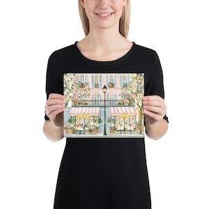 May include: A rectangular art print featuring a watercolor illustration of a Parisian-style street scene with a pink building, striped awnings, yellow umbrellas, outdoor cafe tables, potted flowers, and a central black street lamp post.