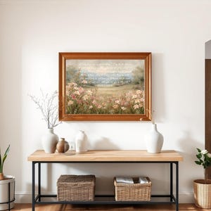May include: A framed landscape painting with a wooden frame hangs above a wooden console table with black metal legs. The painting depicts a field of flowers. Decorative vases and baskets are on the table, and plants are on either side.