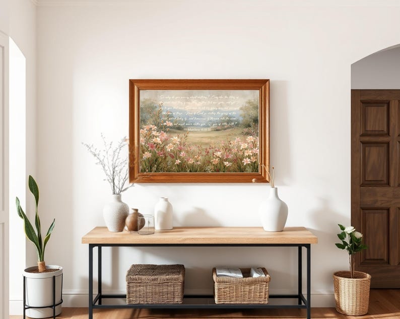 May include: A framed landscape painting with a wooden frame hangs above a wooden console table with black metal legs. The painting depicts a field of flowers. Decorative vases and baskets are on the table, and plants are on either side.