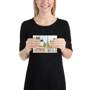 May include: A watercolor print depicting a Parisian street scene with buildings, shops, and the Eiffel Tower in the background. The print features soft colors, including pink, yellow, and green, creating a charming and romantic atmosphere. The print is held by a person.