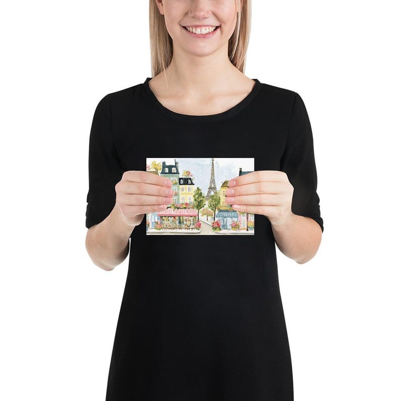 May include: A watercolor print depicting a Parisian street scene with buildings, shops, and the Eiffel Tower in the background. The print features soft colors, including pink, yellow, and green, creating a charming and romantic atmosphere. The print is held by a person.