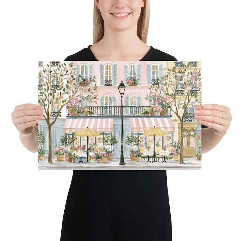 May include: A watercolor-style art print depicting a Parisian-style street scene with a pink building, blue shutters, and two storefronts featuring pink and white striped awnings, yellow umbrellas, potted flowers, and a central black street lamp.
