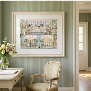 May include: A framed art print depicting a Parisian street scene with a pink building, a black street lamp, and two storefronts with striped awnings and yellow umbrellas. The artwork is displayed in a white mat and a decorative off-white frame.