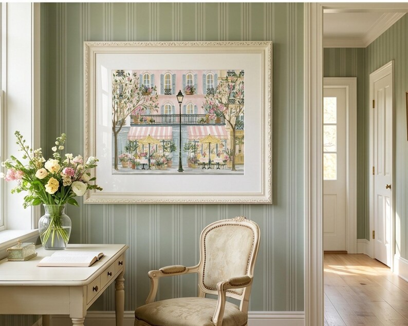May include: A framed art print depicting a Parisian street scene with a pink building, a black street lamp, and two storefronts with striped awnings and yellow umbrellas. The artwork is displayed in a white mat and a decorative off-white frame.