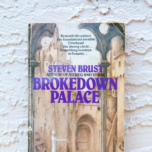 May include: A paperback book cover with a purple title "BROKEDOWN PALACE" by Steven Brust, author of Jhereg and Yendi. The cover features a fantasy illustration of a crumbling building with a figure in a white robe and another figure in a red robe.