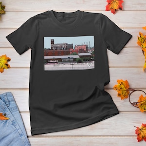Op de afbeelding: Een zwart T-shirt met een vintage foto van een stadsbeeld op de voorkant. De foto toont een groep bakstenen gebouwen met een hoge toren in het midden.