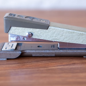 May include: A vintage silver and green stapler with a metal base. The stapler has a textured green top and a silver lever.