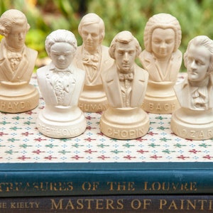 May include: Set of six small white ceramic busts of famous composers, including Haydn, Beethoven, Bach, Mozart, Chopin, and Schumann. The busts are arranged on a stack of books with the titles "Art Treasures of the Louvre" and "Masters of Painting".