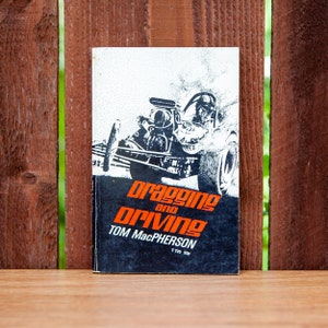 May include: A vintage paperback book titled "Draggin' and Drivin'" by Tom MacPherson. The cover features a black and white illustration of a dragster car with a driver in the cockpit. The book is set against a wooden fence background.