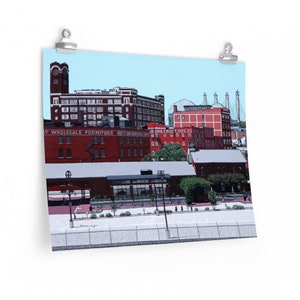 Op de afbeelding: Een digitale illustratie van een stadsbeeld met rode bakstenen gebouwen, een blauwe lucht en een brug op de achtergrond. De tekst "HAY WHOLESALE FURNITURE" is zichtbaar op een van de gebouwen.