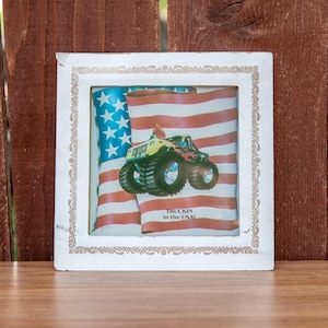 May include: A white framed picture with a monster truck driving on an American flag. The text on the picture reads "Truckin' in the USA!"