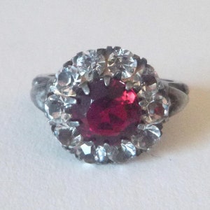 May include: A silver ring with a large red gemstone surrounded by smaller clear gemstones.