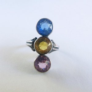 May include: A silver ring featuring three round gemstones in blue, yellow, and pink. The gemstones are stacked vertically, with the blue stone at the top, the yellow in the middle, and the pink at the bottom. The ring has a simple, elegant design.