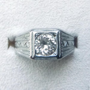 May include: A silver-toned ring with a square face, featuring a large, round, clear gemstone. The band has a geometric design on the sides. The ring is set against a white background.