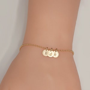 Op de afbeelding: Gouden kettingarmband met drie kleine ronde bedels, elk met een gegraveerde letter. De letters zijn 'a', 'd' en 'e'.
