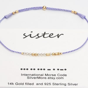 Puede incluir: Un brazalete de cuerda morado con cuentas doradas y plateadas. El brazalete deletrea "hermana" en código Morse internacional.