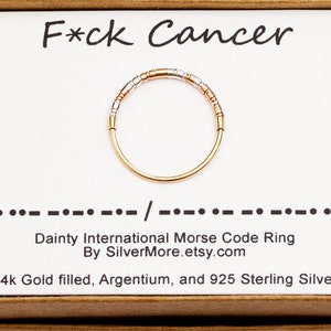Könnte beinhalten: Ein goldfarbener Ring mit Morsecode-Design, präsentiert auf einer Karte mit der Aufschrift "F*ck Cancer". Der Ring hat ein Band mit abwechselnd goldenen und silbernen Perlen. Die Karte enthält auch den Text "Dainty International Morse Code Ring".