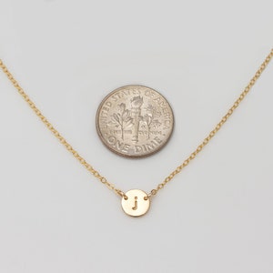 May include: A gold chain necklace with a small gold disc pendant featuring the letter "J". The necklace is shown next to a US dime for size comparison.