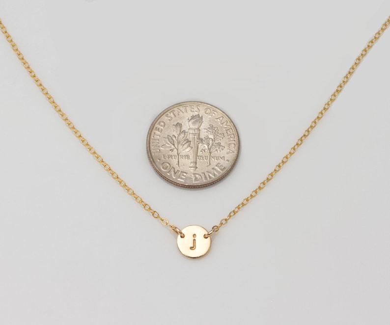 May include: A gold chain necklace with a small gold disc pendant featuring the letter "J". The necklace is shown next to a US dime for size comparison.