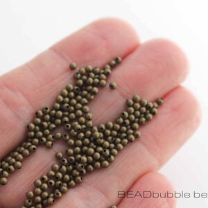 May include: A close-up of a hand holding a large quantity of small, round, bronze-colored beads. The beads are scattered and overlapping, creating a textured and metallic look.