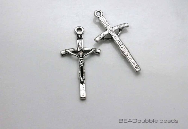 May include: Two silver metal cross charms. One cross charm features a detailed depiction of Jesus Christ on the cross. The other cross charm is a simple, plain cross.