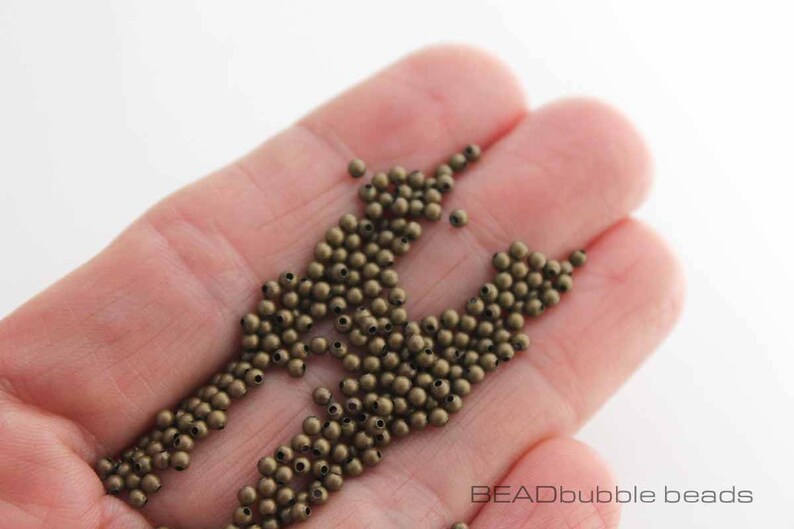 May include: A close-up of a hand holding a large quantity of small, round, bronze-colored beads. The beads are scattered and overlapping, creating a textured and metallic look.