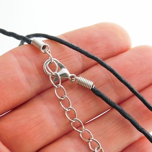 Könnte beinhalten: Eine schwarze Kordelhalskette mit einem silberfarbenen Karabinerverschluss aus Metall, einer kurzen Gliederkette zur Längenverstellung und zwei silberfarbenen, spiralförmigen Endkappen, die die Kordel an den Metallteilen befestigen.