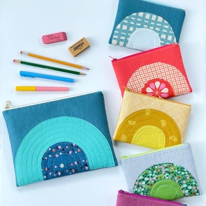 May include: A collection of colorful, quilted zipper pouches with rainbow designs. The pouches are in shades of blue, red, yellow, and green, with various patterned fabrics. Pencils, pens, and erasers are scattered around the pouches.