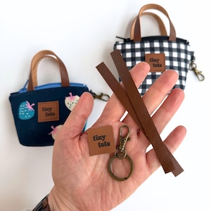 May include: Miniature tote bags in navy blue with strawberry print and black and white gingham patterns. Each bag has brown leather handles and a small tag that says "tiny tote". Accessories include a key ring, clasp, and leather strips.