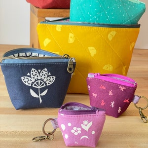 May include: Five zippered pouches in various colours and patterns. The pouches are made of fabric and have a quilted design. The pouches are arranged in a pyramid shape, with the largest pouch at the bottom and the smallest pouch at the top.