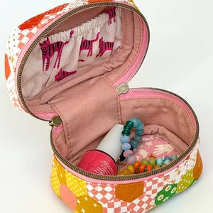 以下が含まれることがあります： カラフルなパッチワークデザインの、開いたキルティングコスメティックバッグ。内側はピンクの裏地とピンクのゼブラ柄のポケットが付いています。バッグにはマニキュアのボトルとビーズのジュエリーが入っています。