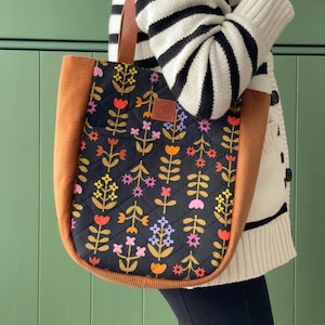 May include: A brown and black tote bag with a floral pattern. The bag has a brown corduroy trim and a brown leather handle.