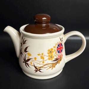 May include: Cream ceramic teapot with a brown lid and handle. The teapot has a floral design with yellow flowers and brown branches. A badge with the Union Jack flag is on the side. The teapot is designed for brewing tea.