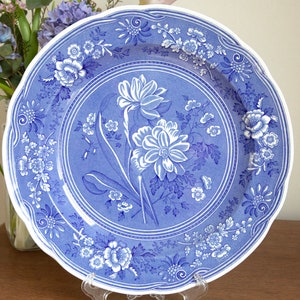 May include: A blue and white ceramic dinner plate with a floral design. The plate features a central motif of stylised flowers with a blue background and white accents. The rim of the plate is decorated with a repeating pattern of smaller flowers.