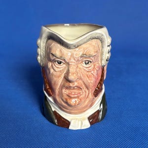 May include: A ceramic figurine of a person wearing a white wig and a black robe. The figurine is a mug with a detailed face and a brown and white patterned robe.