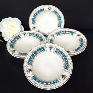 May include: A set of four white porcelain bowls with a teal and gold floral design. The bowls have a scalloped edge and are arranged on a black surface. A white rose is in the background.