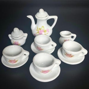 May include: A miniature white porcelain tea set. The set includes a teapot, sugar bowl, and four cups with saucers. Each piece is decorated with pink rose floral accents. The tea set is designed for children's play or display.