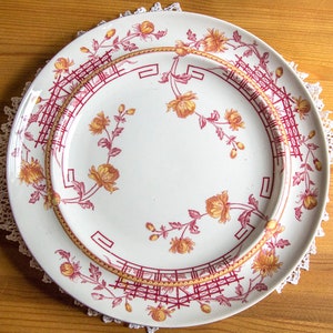 May include: A white ceramic dinner plate with a red and gold floral pattern. The plate has a raised rim and a scalloped edge.