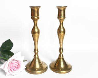 May include: A pair of vintage brass candlesticks with a classic design. The candlesticks have a flared base, a slender, curved stem, and a cup-shaped top. A pink rose and green leaves are in the background.