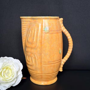 May include: A vintage, orange-yellow ceramic pitcher with a textured surface and a looped handle. The pitcher features a decorative design on the front and is accented with small, light green details. A white rose is in the foreground.