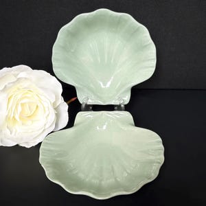 May include: Two pale green seashell-shaped ceramic dishes. The dishes are stacked on top of each other. A large white rose with yellow center is to the left of the dishes. The background is black.