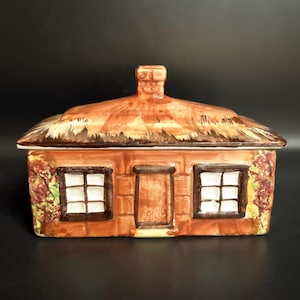 May include: A ceramic cottage-shaped trinket box with a thatched roof and brick-like walls. The windows and door are outlined in brown, with white window panes. The box has a chimney and is painted in shades of brown and orange.