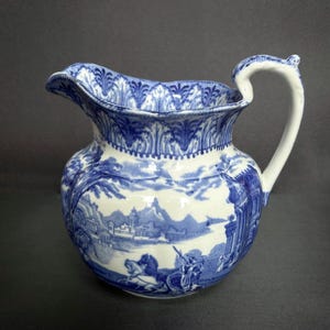 May include: A vintage blue and white ceramic pitcher with a detailed landscape scene. The pitcher features a curved handle and a scalloped rim. The design includes buildings, figures, and trees, with a decorative border around the top.