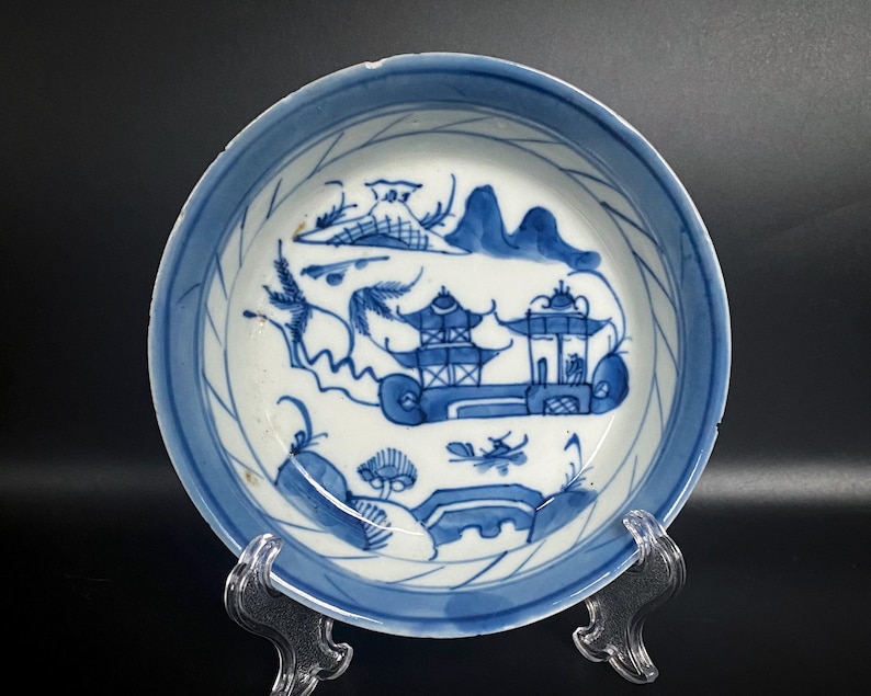 Puede incluir: Un plato de porcelana azul y blanco con un dise&ntilde;o decorativo de un paisaje chino con una pagoda y &aacute;rboles. El plato tiene un fondo blanco con detalles azules y un borde azul.
