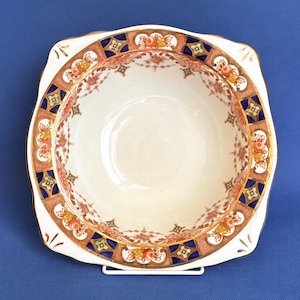 May include: A white, square-shaped bowl with a decorative rim featuring orange, blue, and gold floral patterns. The bowl has a scalloped edge and a gold trim. The interior is plain white. The bowl is likely made of porcelain or ceramic.