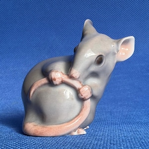 May include: A ceramic figurine of a mouse in a curled position. The mouse is primarily gray with pink accents on its ears and tail. The figurine is set against a blue background.