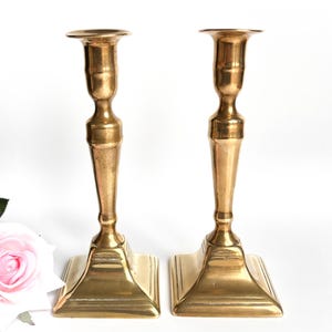 Peut inclure: Deux bougeoirs en laiton vintage avec des bases carrées et un design classique. Les bougeoirs ont une teinte dorée chaude et sont présentés sur un fond blanc. Une rose rose est visible dans le coin inférieur gauche.