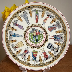 Peut inclure: Une assiette en céramique blanche avec un bord rouge présente un motif circulaire de personnages dans différents styles vestimentaires à travers l'histoire. L'assiette est intitulée "Fashion Through The Ages 11th Century to 20th Century".