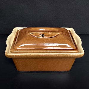 May include: A brown ceramic casserole dish with a lid. The lid features a handle and a small vent hole. The dish rests on a lighter-coloured base with curved edges. The design is simple and classic, suitable for various culinary uses.